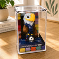 FIFA World Cup figurine in a clear display case on a wooden surface with books and a plant in the background.
